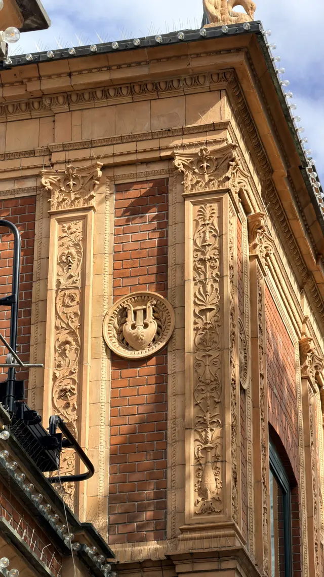 Et øvre hjørne af en pavillon: en rund medaljon med en lyre mellem felter af rød mursten og terrakottapilastre med blomster, under gesimsen.