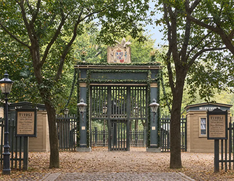Tivolis tremmeport mellem træer, med skilte for en udstilling på hver side, et plankeværk langs gaden og et lille billetkontor til højre.