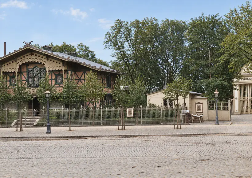 Til venstre en bindingsværkspavillon med udskårne vindskeder bag et jerngelænder med Tivoli-plakater; til højre en lille hvid kontorbygning og porten. Gaslygter, unge træer og skinner i brostenene.