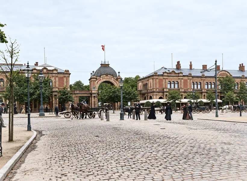 Porten og begge pavilloner set på afstand over en bred brostensgade, med hestevogne, fodgængere og gaslygter; der er endnu ingen elektriske pærer på porten.